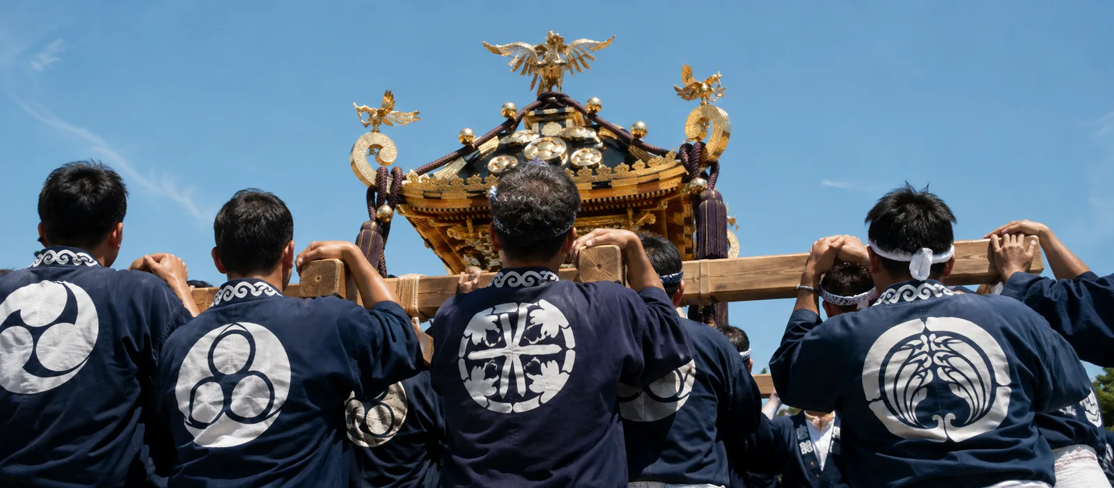 揃いの半纏を着た担ぎ手が神輿を担ぐ場面。背の会名と紋が見える構図