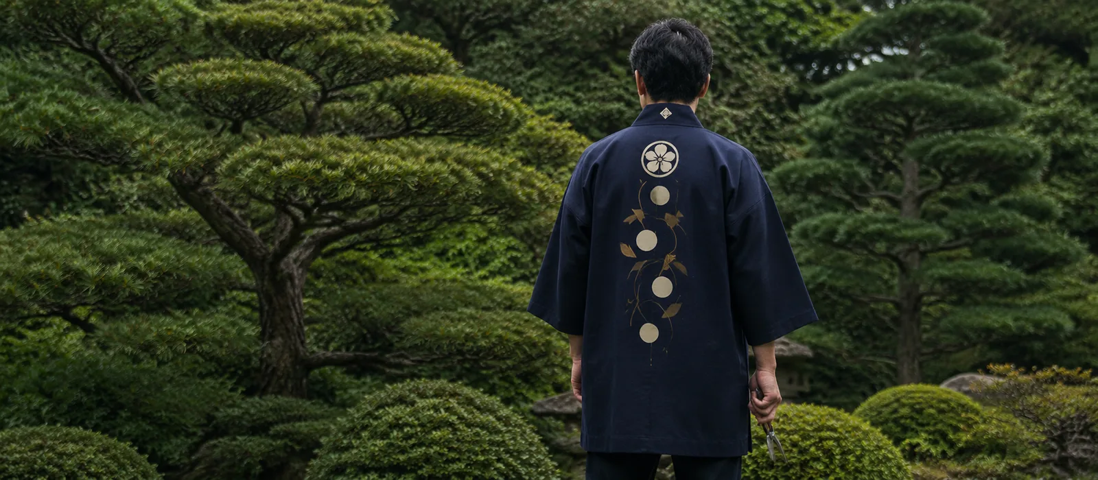 庭木の前に立つ、屋号を背に入れた半纏姿の植木屋。背面が見える構図